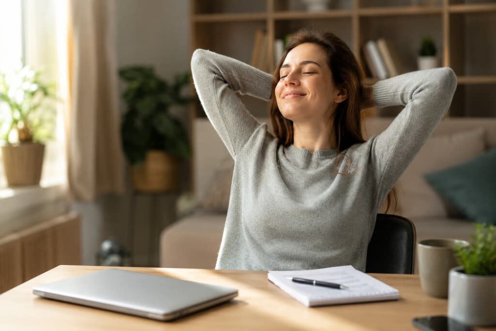 Estudiante universitaria online sonriendo con satisfacción después de completar una tarea académica en casa, sentada frente a un portátil cerrado y apuntes terminados.